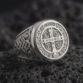 Load image into Gallery viewer, White gold Saint Benedict signet ring resting on black volcanic stone, showcasing the engraved Catholic cross medal, traditional Latin letters, PAX symbol, intricate Celtic-inspired shoulder patterns, and bright polished silver finish.