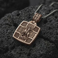 Load image into Gallery viewer, Bronze Saint George Slaying Dragon pendant on black volcanic rock, featuring the warrior saint on horseback with a spear.
