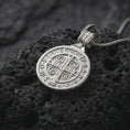 Load image into Gallery viewer, Polished Silver Saint Benedict medal necklace on black volcanic rock, featuring a round religious pendant with cross symbols and detailed lettering.