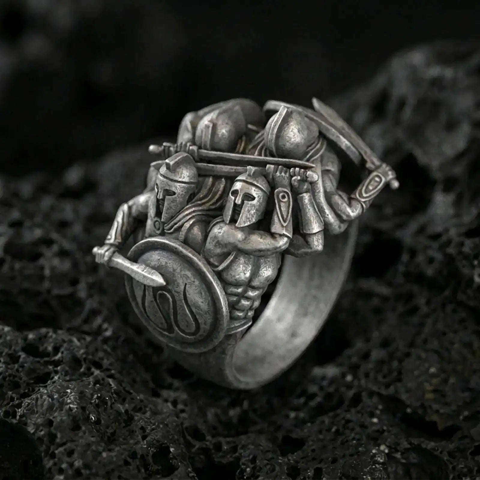 Sterling silver Spartan warrior ring with an oxidized finish, sculptural battle scene featuring armored Greek soldiers, shield and sword detail, macro on black volcanic rock