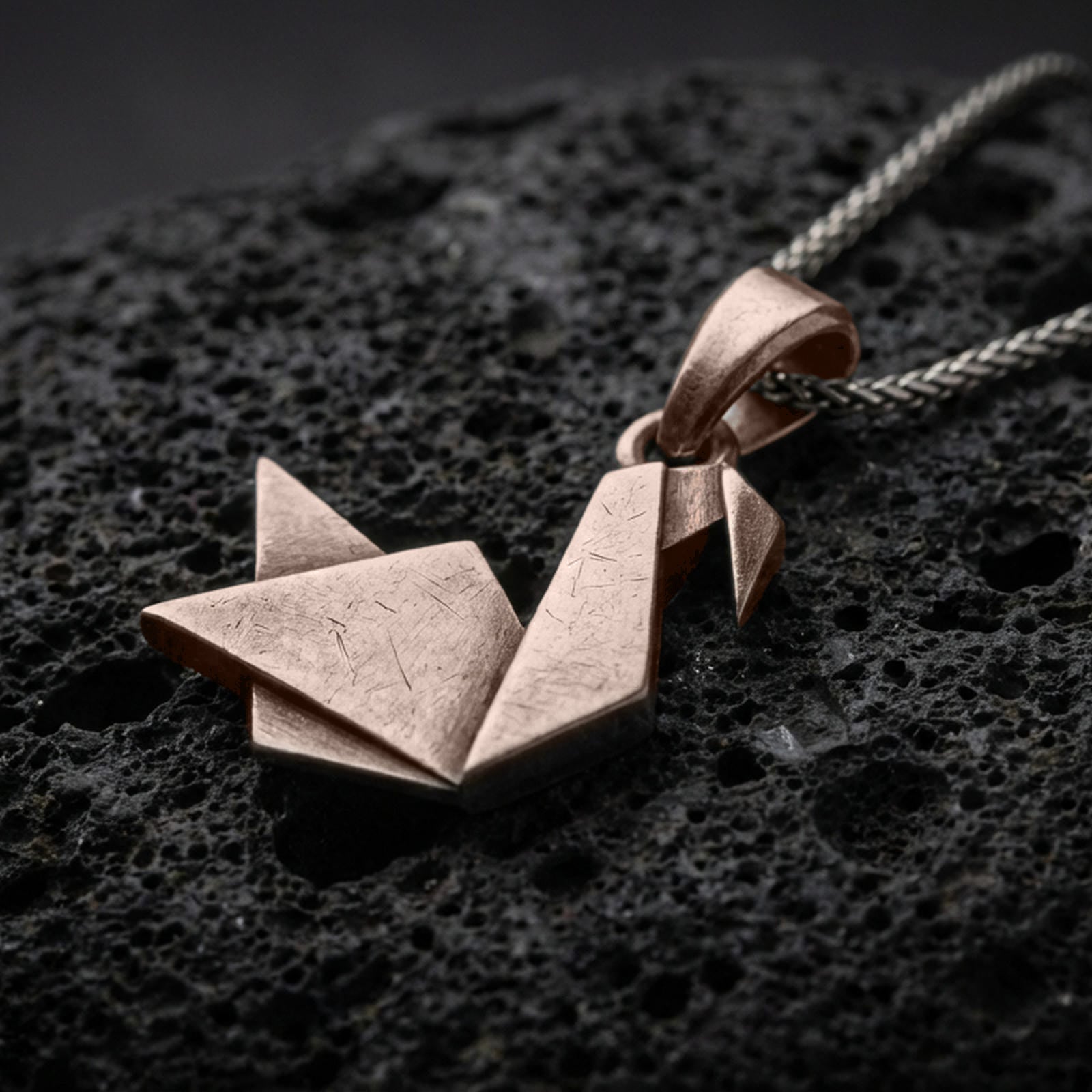 Bronze origami swan pendant with folded geometric silhouette, minimalist angular lines, and elegant bird design displayed on dark volcanic rock in a close-up luxury jewelry composition
