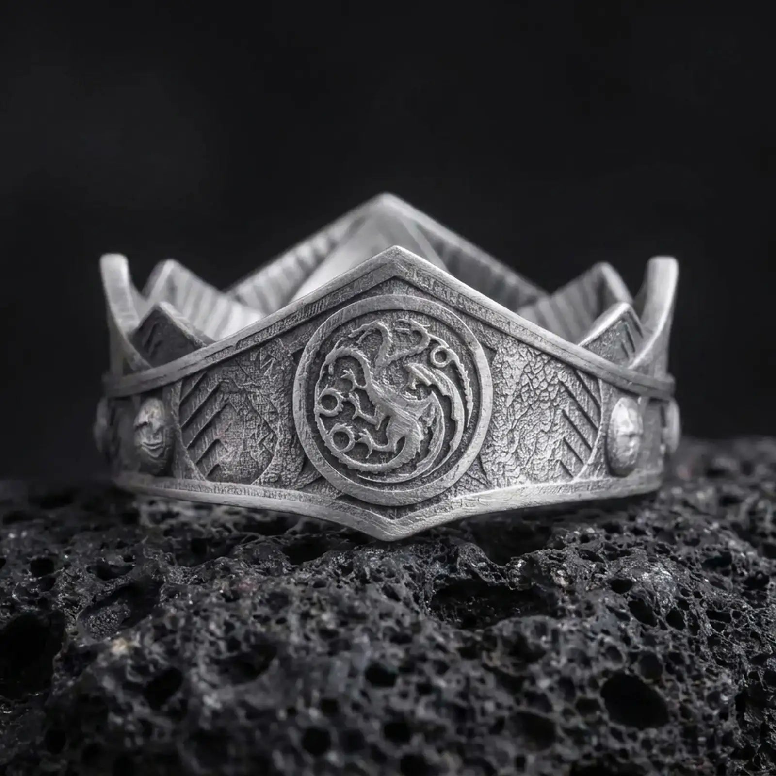 Oxidized Targaryen dragon ring on textured volcanic rock, close-up studio shot highlighting the three-headed dragon emblem, shield-like face, engraved band details, and antique silver contrast.