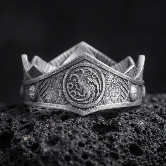 Oxidized Targaryen dragon ring on textured volcanic rock, close-up studio shot highlighting the three-headed dragon emblem, shield-like face, engraved band details, and antique silver contrast.