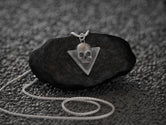 May include: A silver skull pendant necklace with a triangular frame. The pendant is hanging from a silver chain on a black rock.