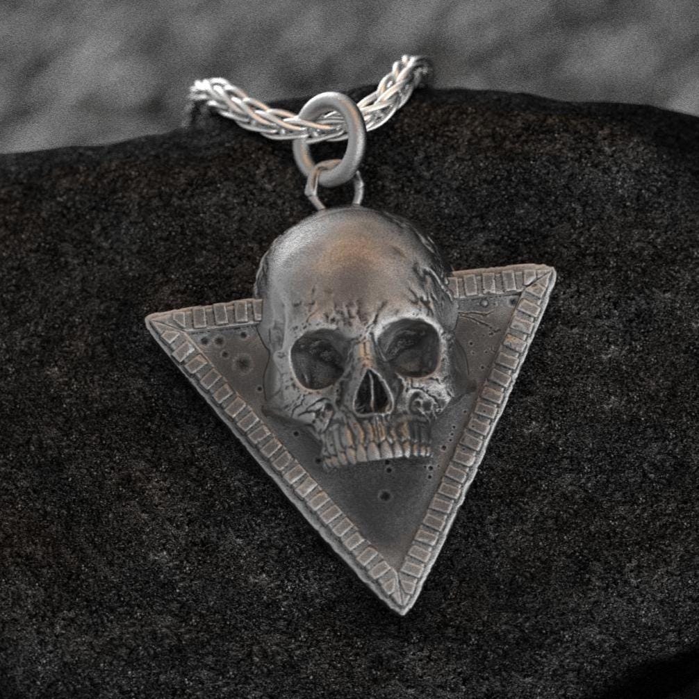May include: A silver skull pendant with a detailed design. The skull is set in a triangular frame with a textured border. The pendant is on a silver chain.
