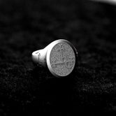 May include: A silver signet ring with a circular, textured top featuring an etched design. The ring has a classic style and is displayed on a dark gray surface. The detailed engraving on the face of the ring is visible.