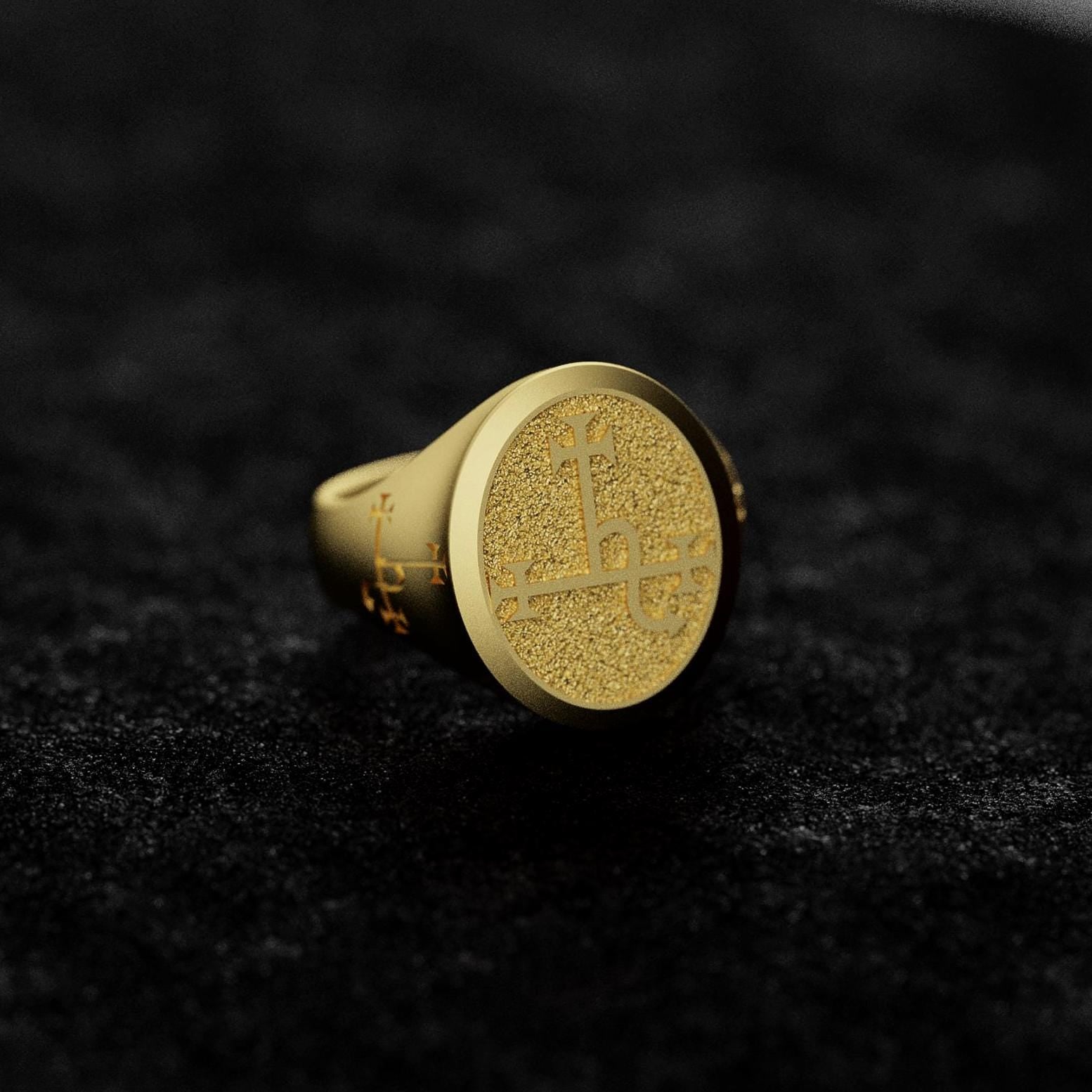 May include: A gold signet ring with a textured, circular top featuring an engraved design. The ring has a classic style and is displayed on a dark gray surface.