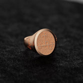 May include: Rose gold signet ring with a textured, circular face featuring an engraved emblem. The ring&#39;s band is visible, and the overall design is simple and elegant. The signet ring is displayed on a dark gray textured surface.