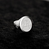 May include: A silver signet ring with a circular, textured top featuring an etched design. The ring has a classic style and is displayed on a dark gray surface. The detailed engraving on the face of the ring is visible.