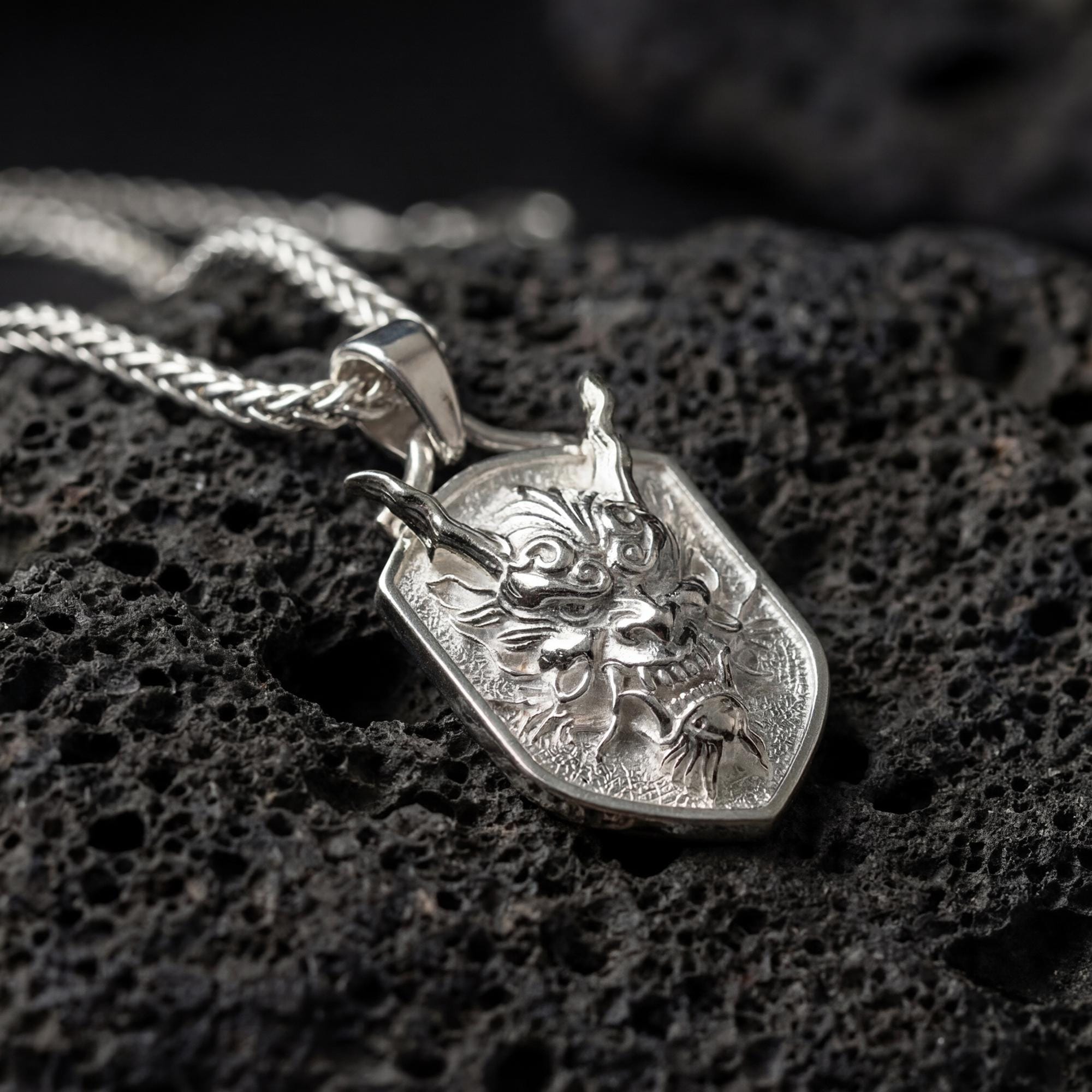 Polished silver oni mask dragon dog tag necklace displayed on volcanic stone background with sharp sculpted relief