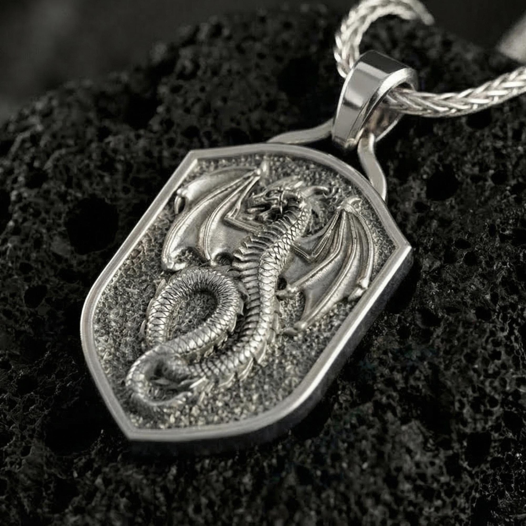 Polished silver coiled dragon shield necklace displayed on volcanic stone background with sharp sculpted relief