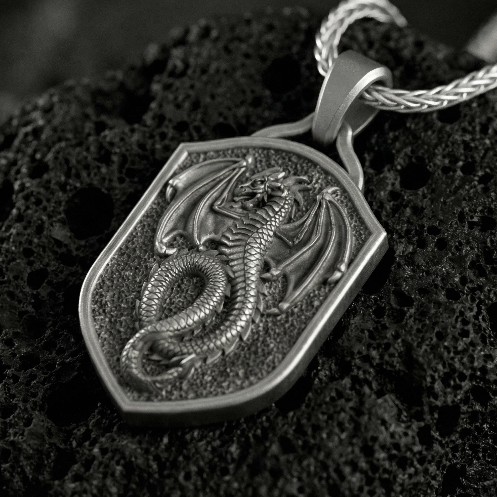 Oxidized coiled dragon shield pendant necklace photographed on volcanic rock with deeply engraved fantasy dragon detail