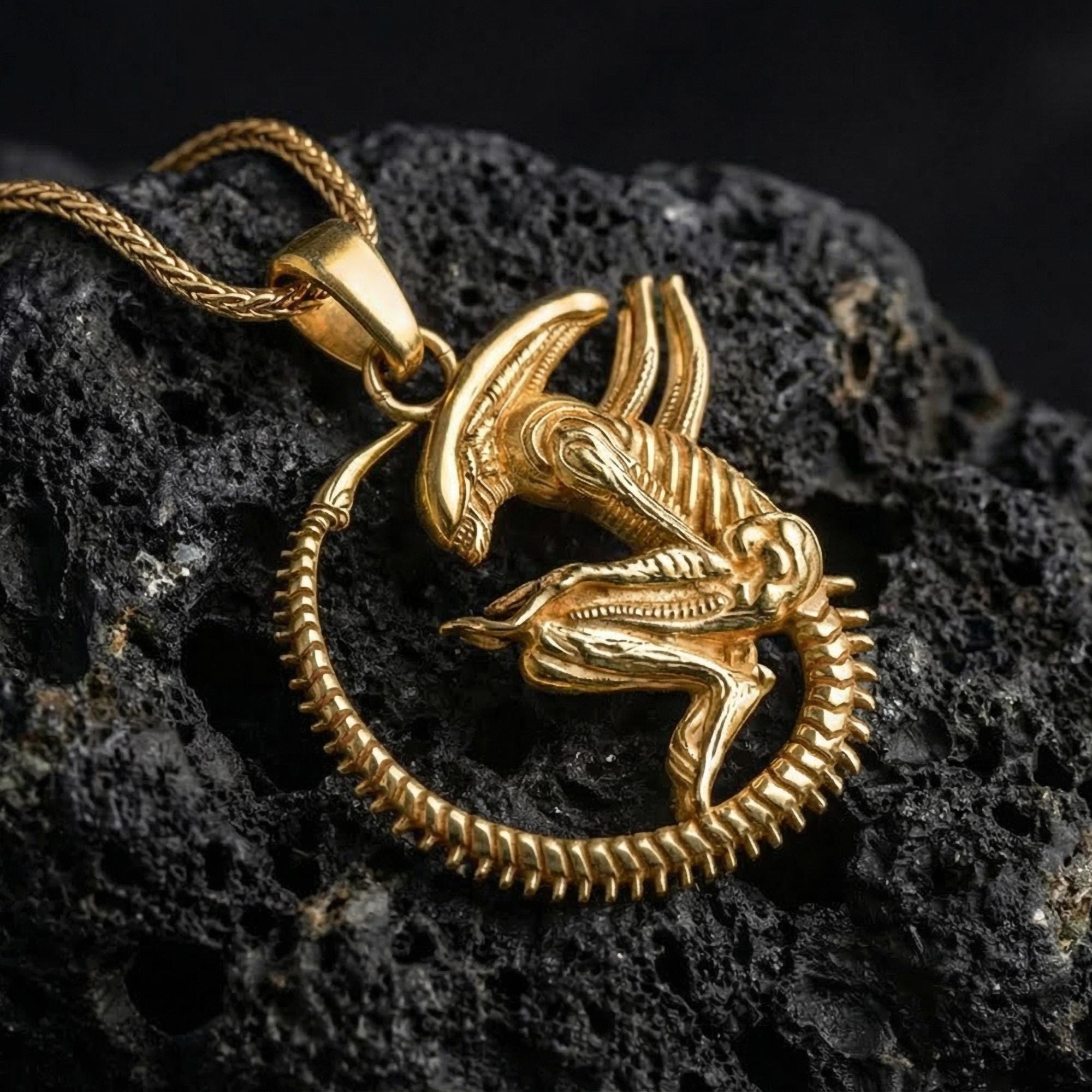 Gold plated alien xenomorph pendant showcased on volcanic rock as dark sci-fi horror statement jewelry