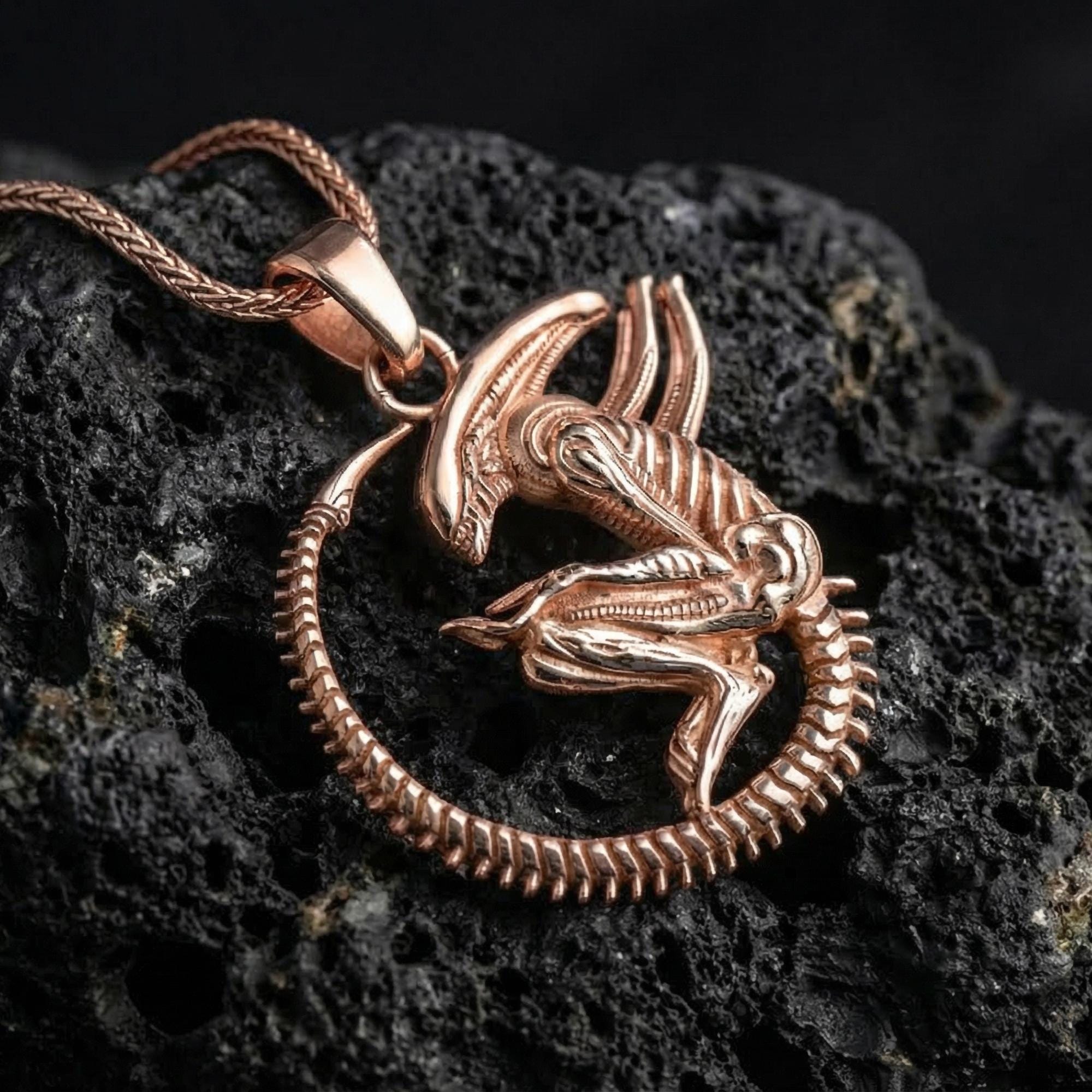 Rose gold plated xenomorph creature pendant necklace presented on volcanic rock surface with futuristic horror aesthetic