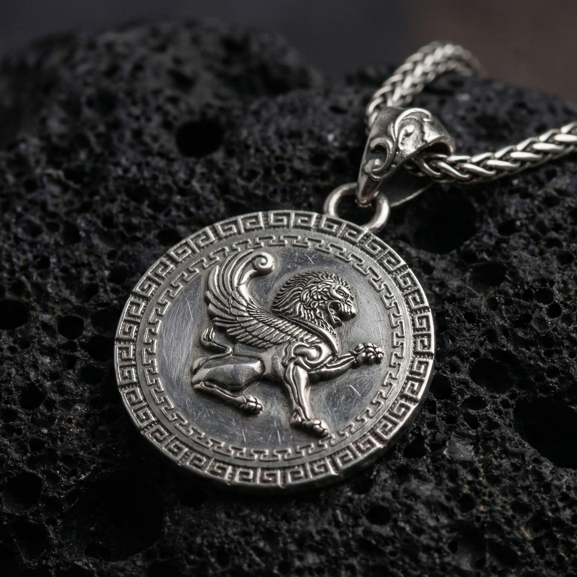 Oxidized winged lion pendant necklace photographed on volcanic rock with detailed mythical griffin style relief and Greek key border