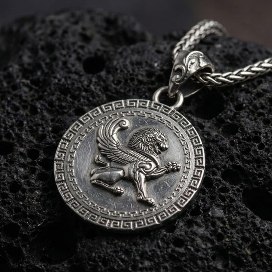 Oxidized winged lion pendant necklace photographed on volcanic rock with detailed mythical griffin style relief and Greek key border