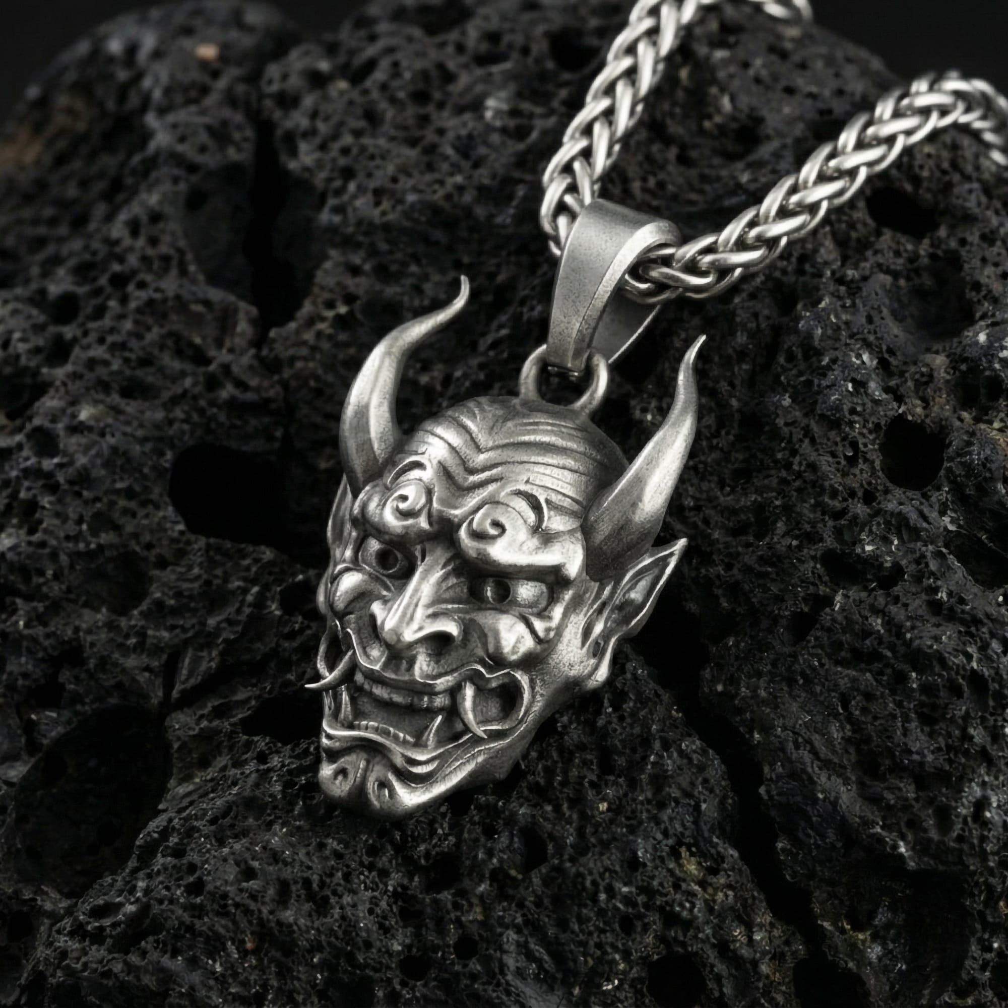 Oxidized oni mask pendant necklace photographed on volcanic rock with Japanese demon face relief and sharp horn detail