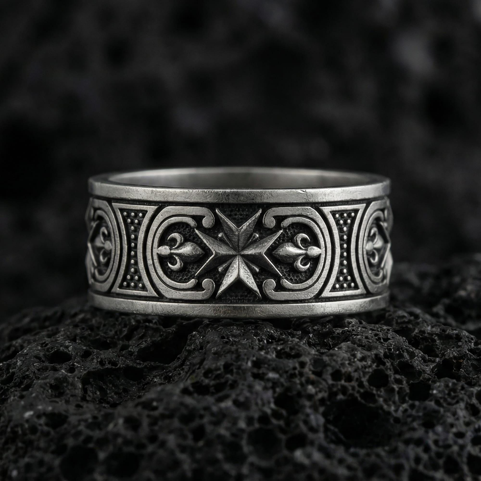 Oxidized Maltese cross Knights Hospitaller band ring photographed on volcanic rock, featuring crisp star-cross emblem and fleur de lis scrollwork detail