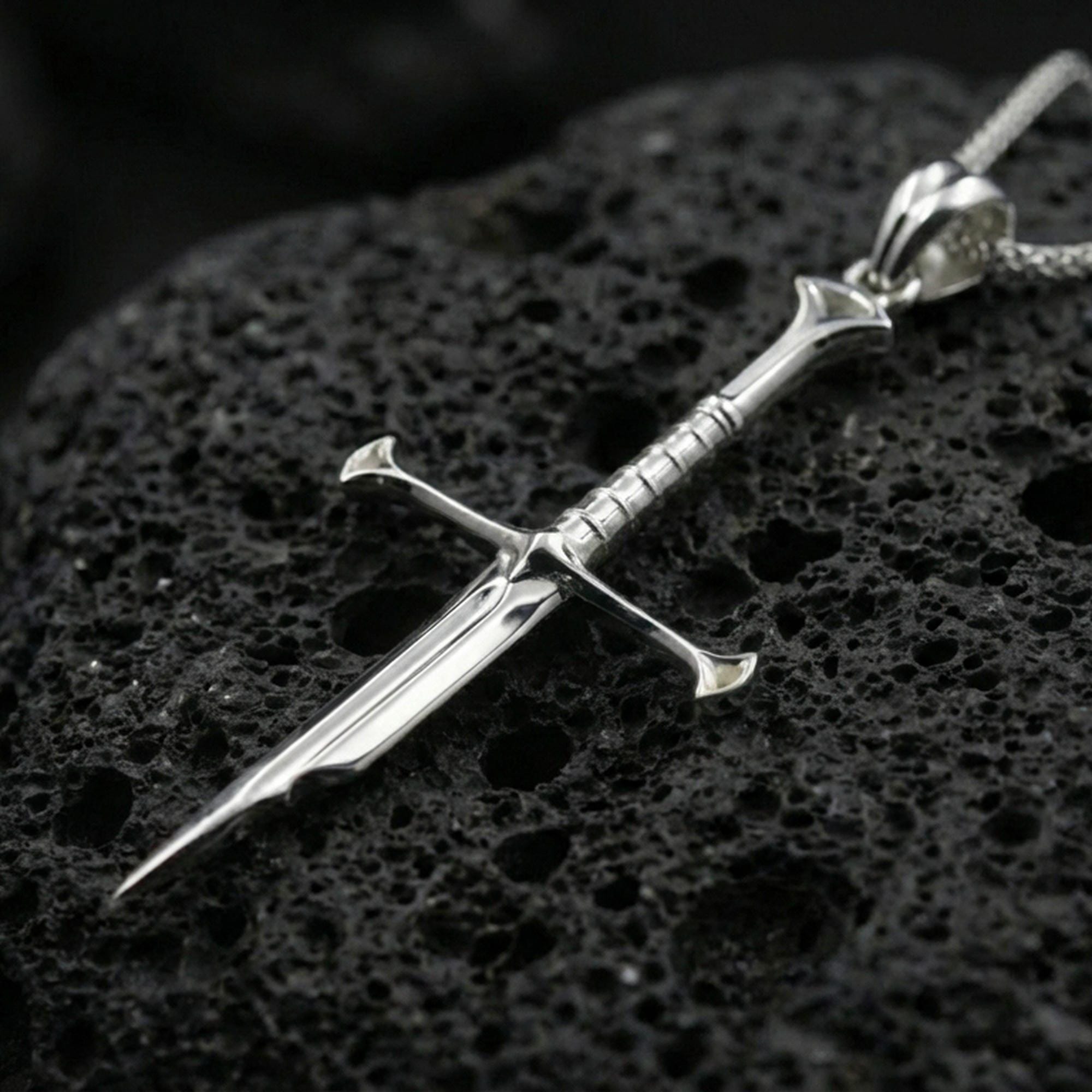 Polished silver sword necklace pendant on black volcanic stone background, clean reflections, crisp crossguard and edge