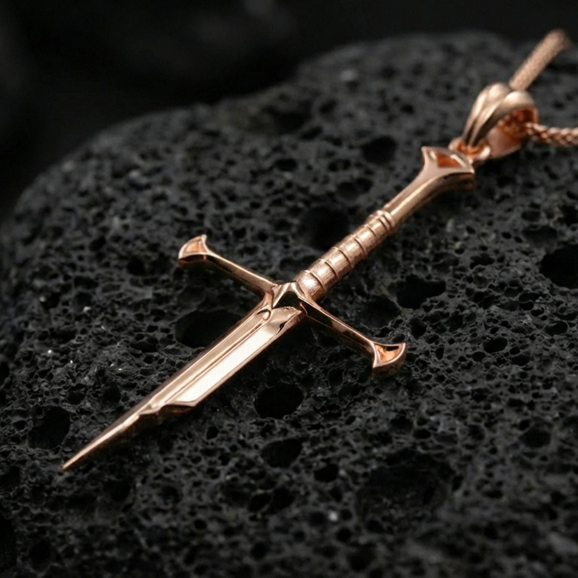 Rose gold plated sword jewelry pendant on volcanic rock, premium macro product photo, detailed hilt and blade profile