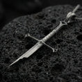 Load image into Gallery viewer, Oxidized sword pendant on textured volcanic rock, macro close-up, dark premium studio lighting, sharp blade detailing
