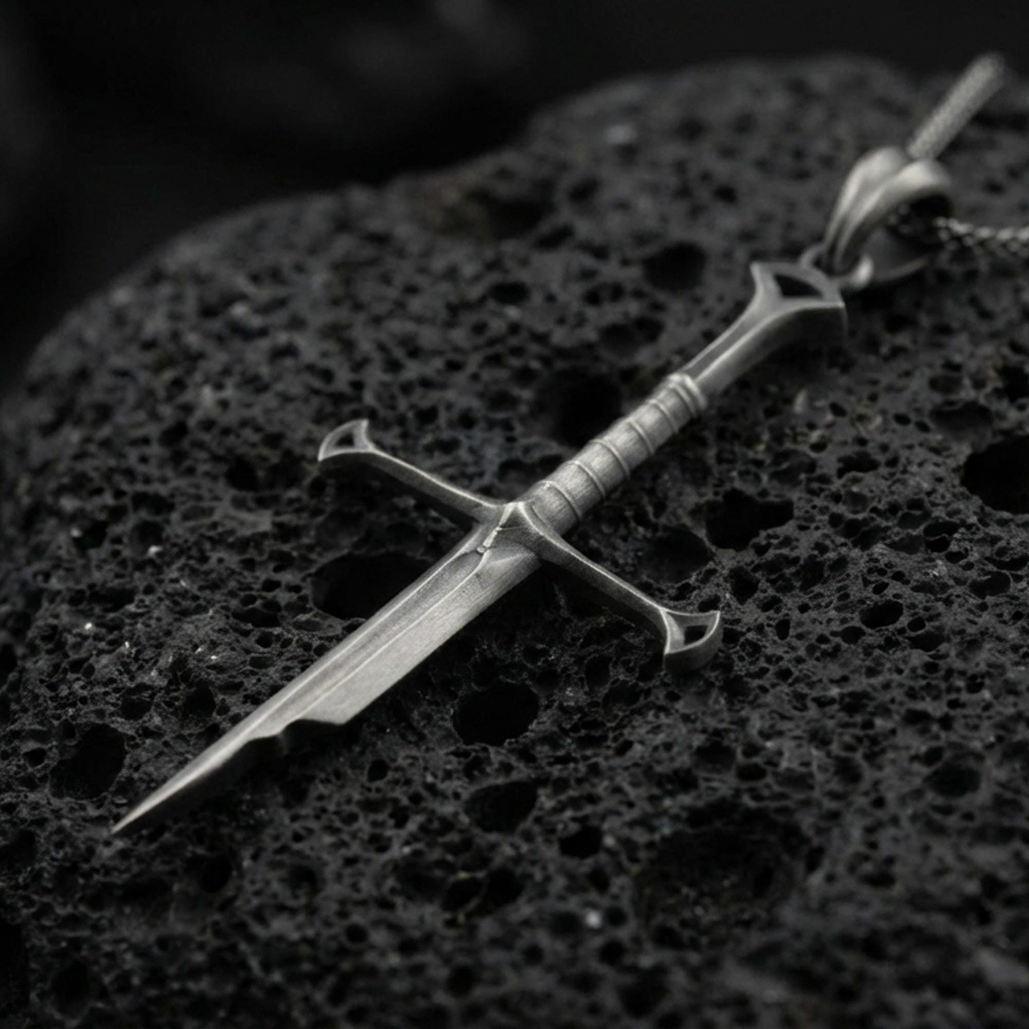 Oxidized sword pendant on textured volcanic rock, macro close-up, dark premium studio lighting, sharp blade detailing