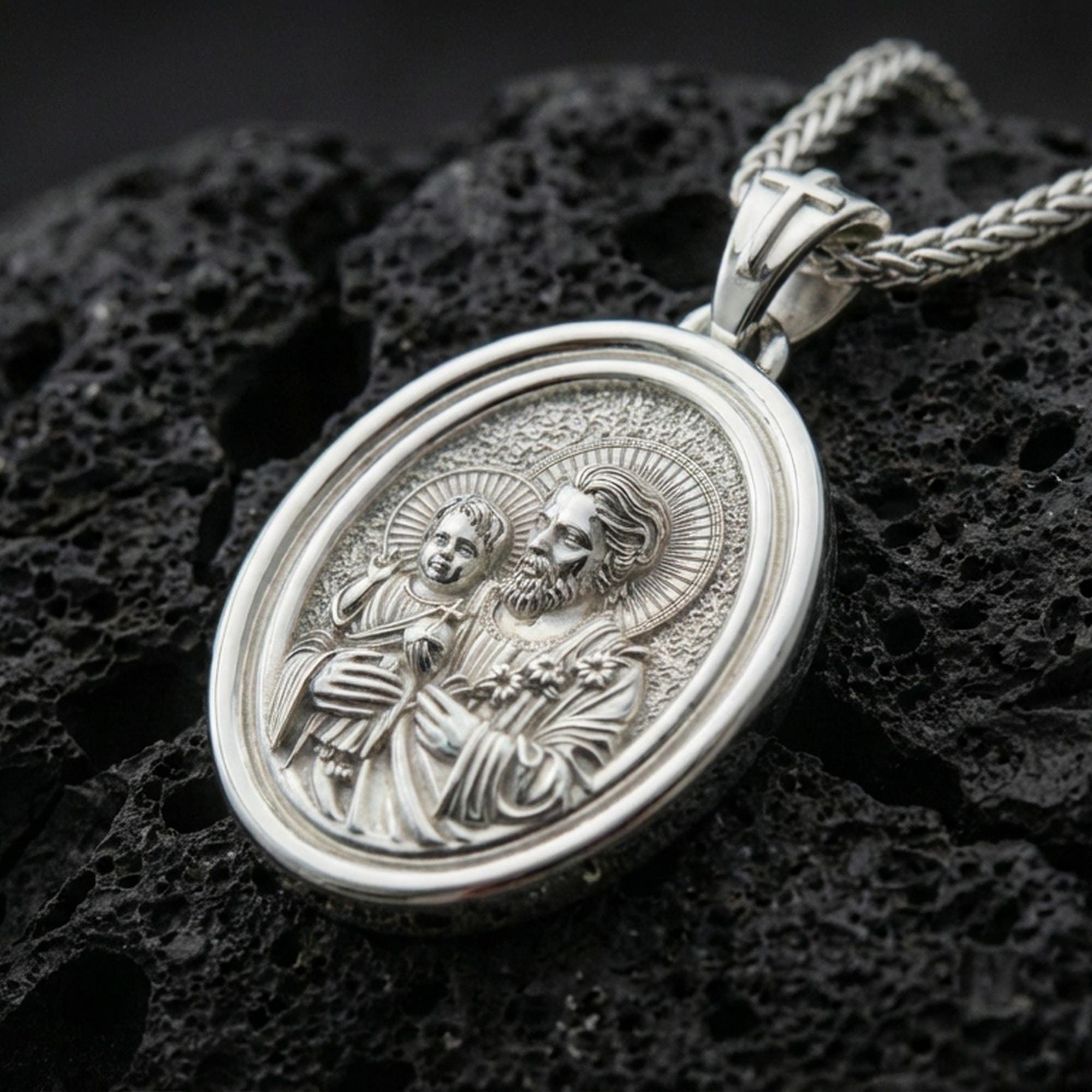 Polished silver Saint Joseph necklace pendant on black volcanic stone background, crisp relief of Saint Joseph and child, high-end product shot