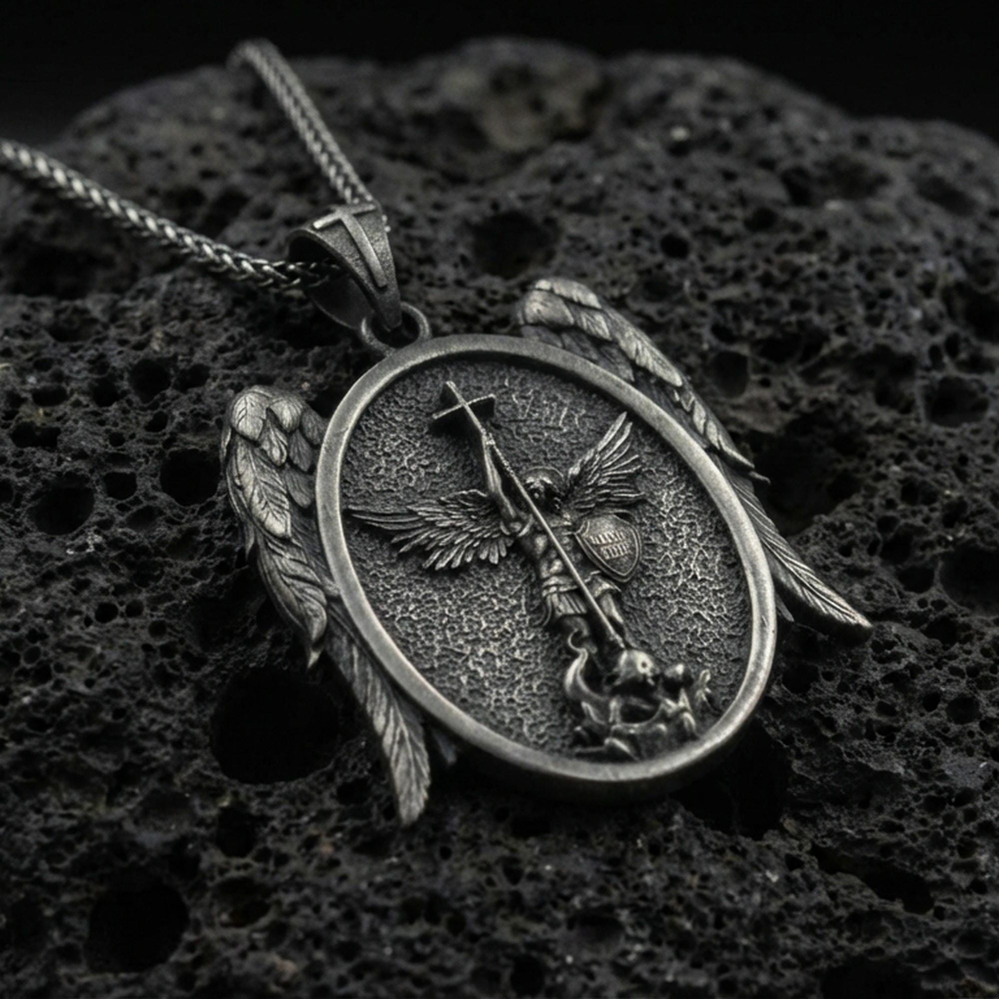 Oxidized Saint Michael archangel pendant on textured volcanic rock, macro close-up, oval medallion with wing accents, premium studio lighting