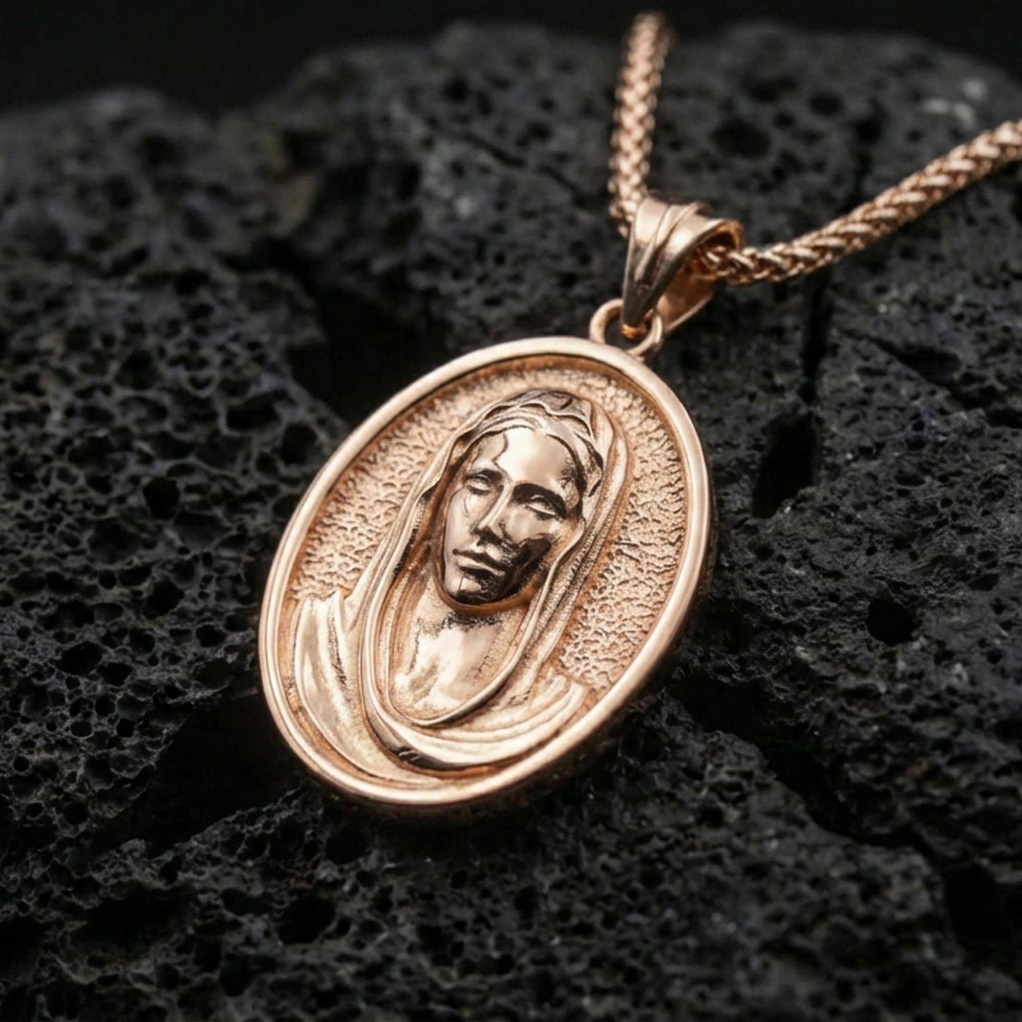 Rose gold plated Veiled Madonna religious pendant on volcanic rock, premium macro product photo, sharp oval silhouette and veil detailing