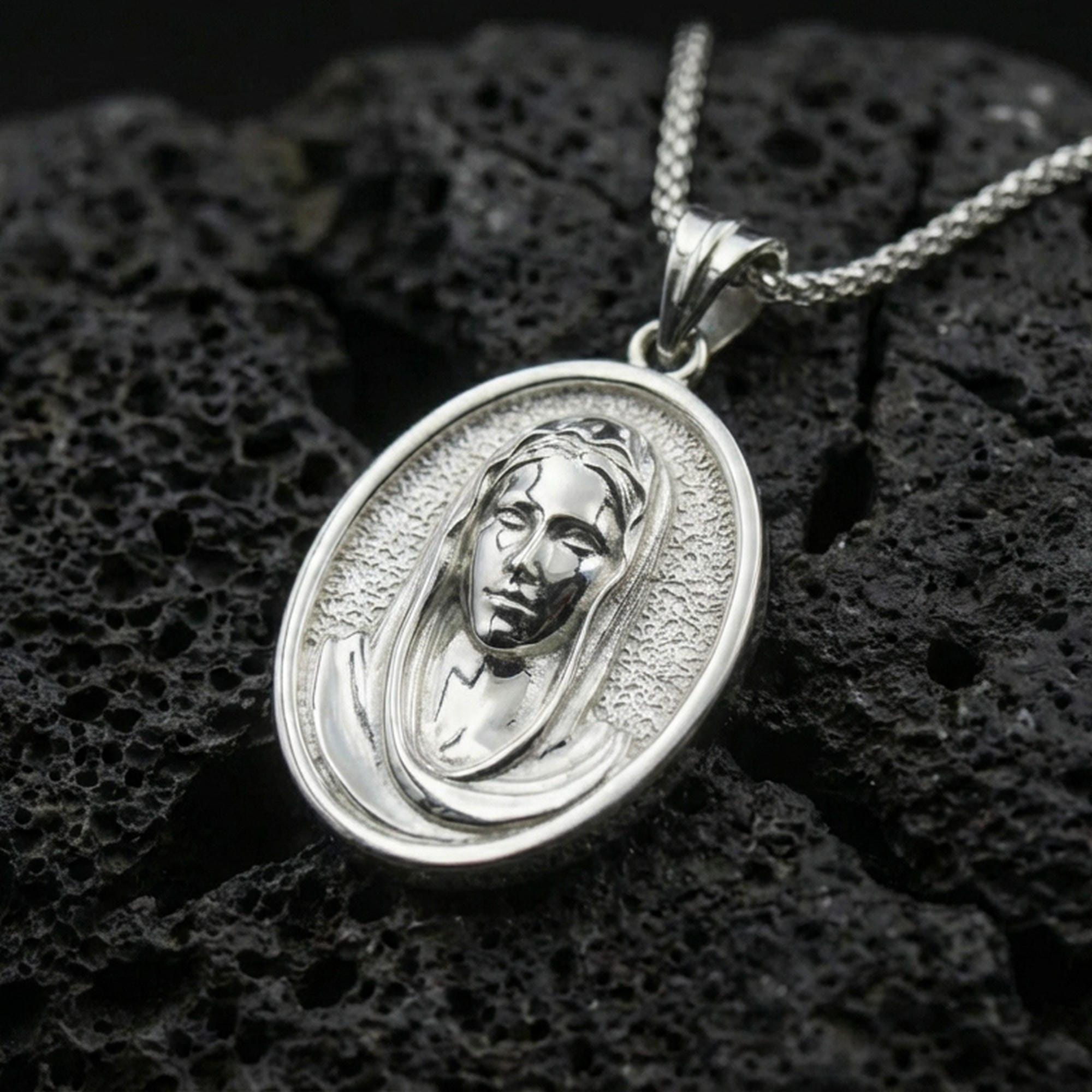 Polished silver Veiled Madonna necklace pendant on black volcanic stone background, crisp oval frame and veil texture, high-end product shot