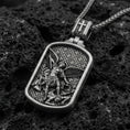 Load image into Gallery viewer, Oxidized Archangel Michael dog tag pendant on textured volcanic rock, macro close-up, angel defeating demon relief, premium studio lighting
