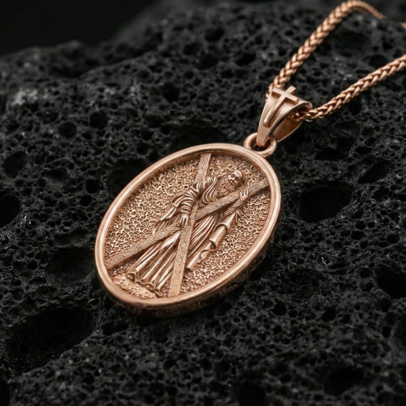Rose gold plated Saint Andrew oval medallion pendant on volcanic rock surface, macro product shot, clean silhouette and sharp engraving