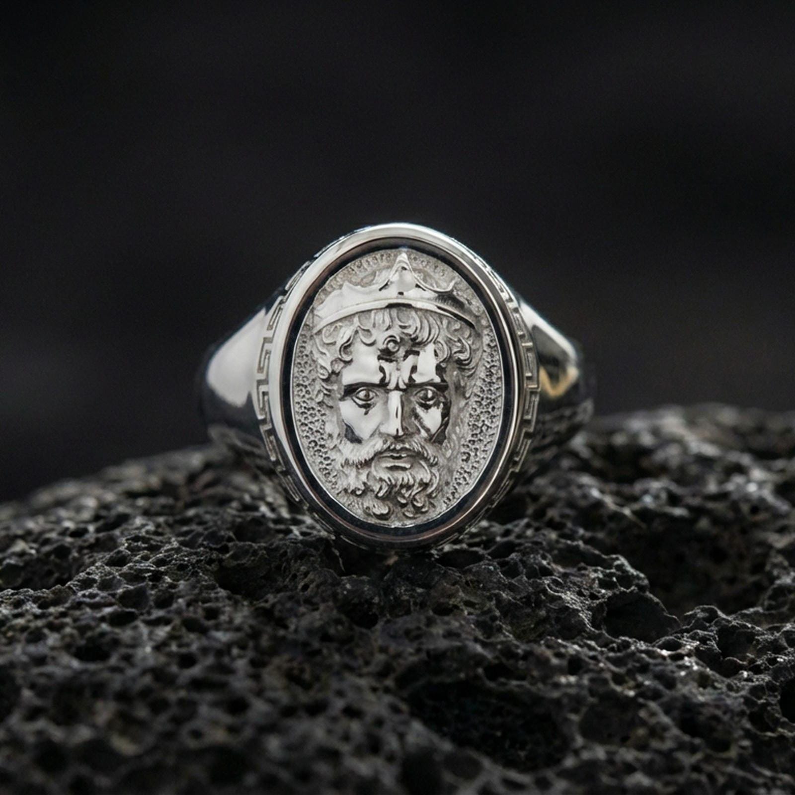 Polished silver Poseidon signet ring on black volcanic stone, Neptune-inspired crowned face relief, Greek key side detail, close-up
