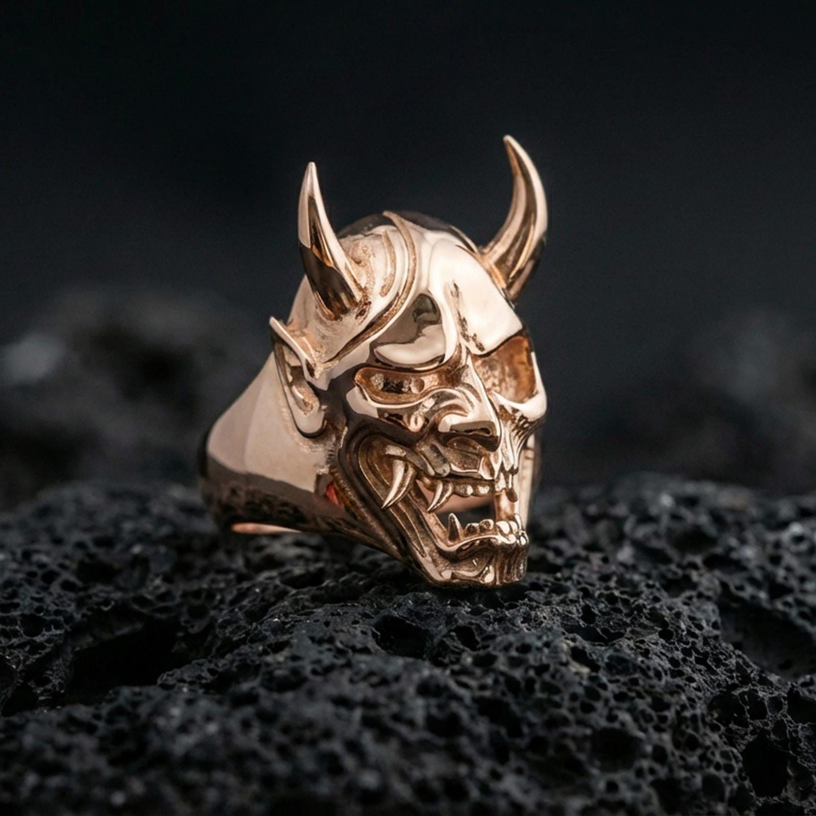 Oni mask skull ring in a rose gold plated finish on volcanic rock, ornate yokai demon face with horned profile, luxury studio close-up