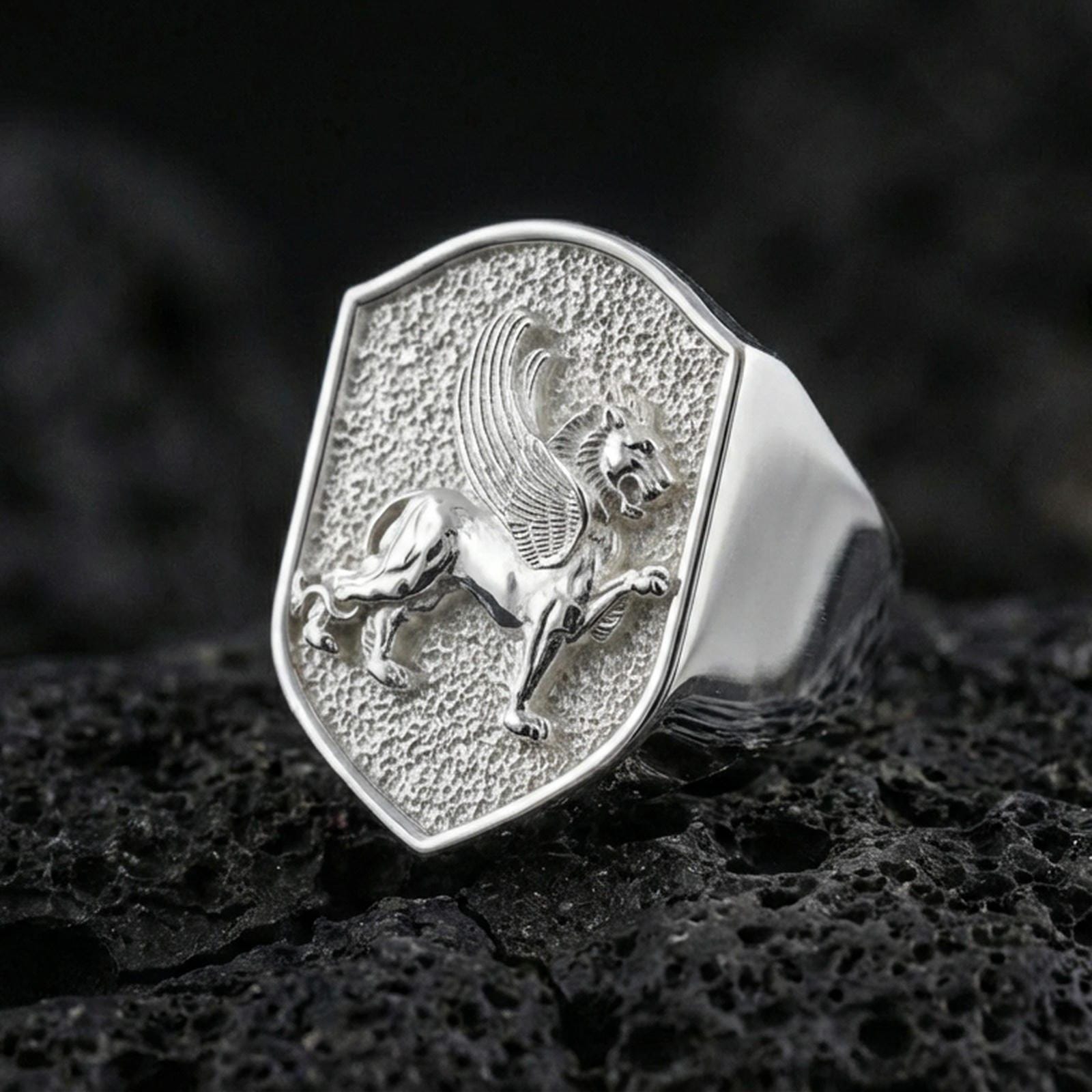 Sterling silver griffin ring in polished silver finish on volcanic rock, detailed winged gryphon engraving with textured background, close-up photo