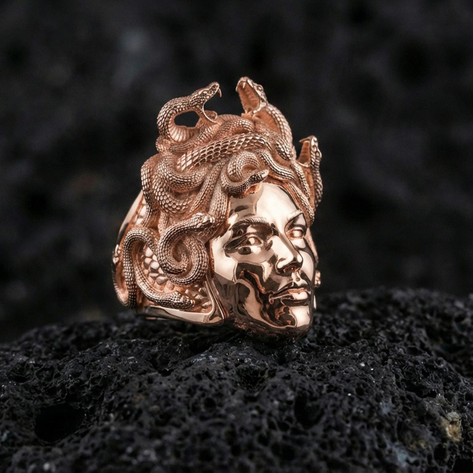 Medusa ring in a rose gold plated finish on volcanic rock, ornate Greek mythology gorgon portrait with snakes, luxury studio close-up