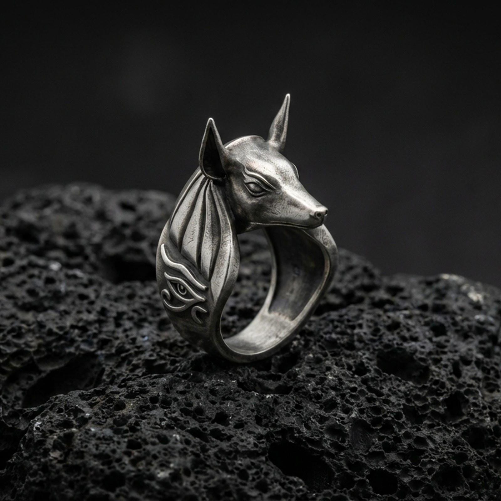 Sterling silver Anubis ring in an oxidized finish on volcanic rock, jackal-headed Egyptian god with Eye of Horus side engraving, dramatic macro product shot