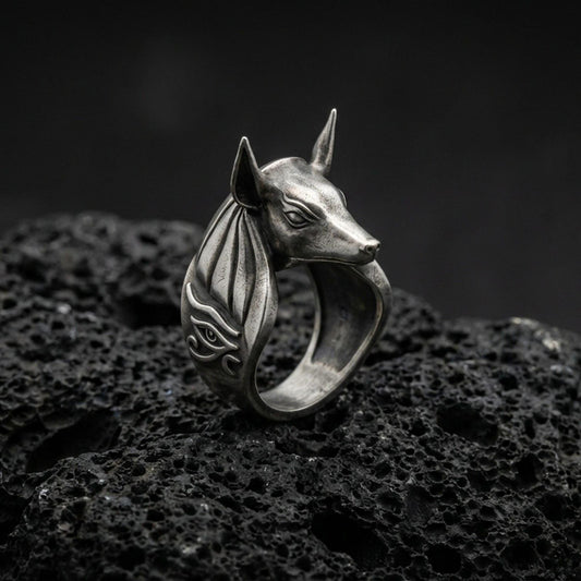 Sterling silver Anubis ring in an oxidized finish on volcanic rock, jackal-headed Egyptian god with Eye of Horus side engraving, dramatic macro product shot