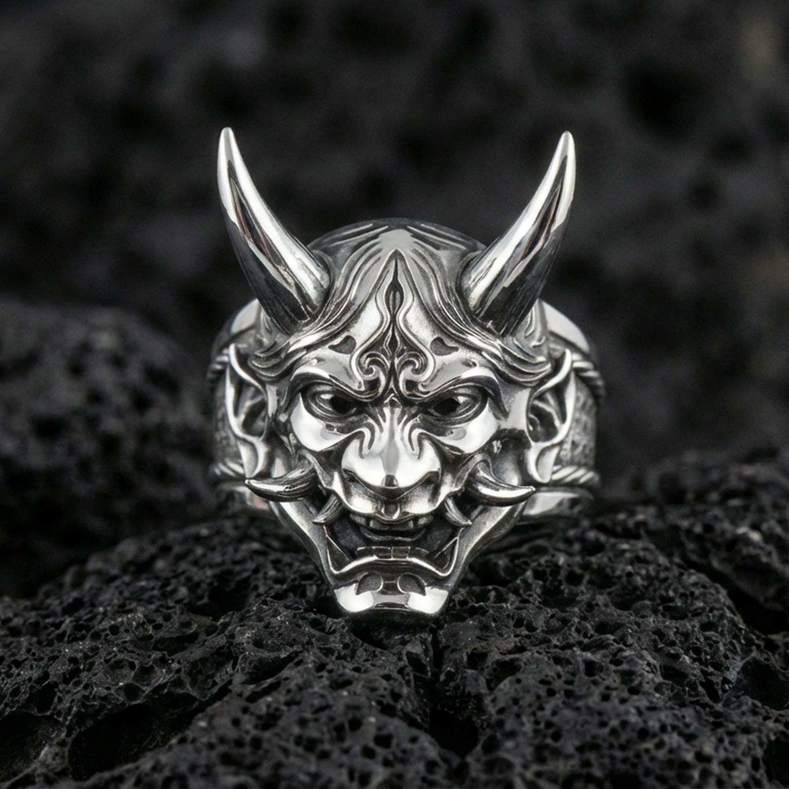 Oni demon mask ring in polished silver finish on volcanic rock, raised samurai-inspired yokai faceplate, premium close-up jewelry photo