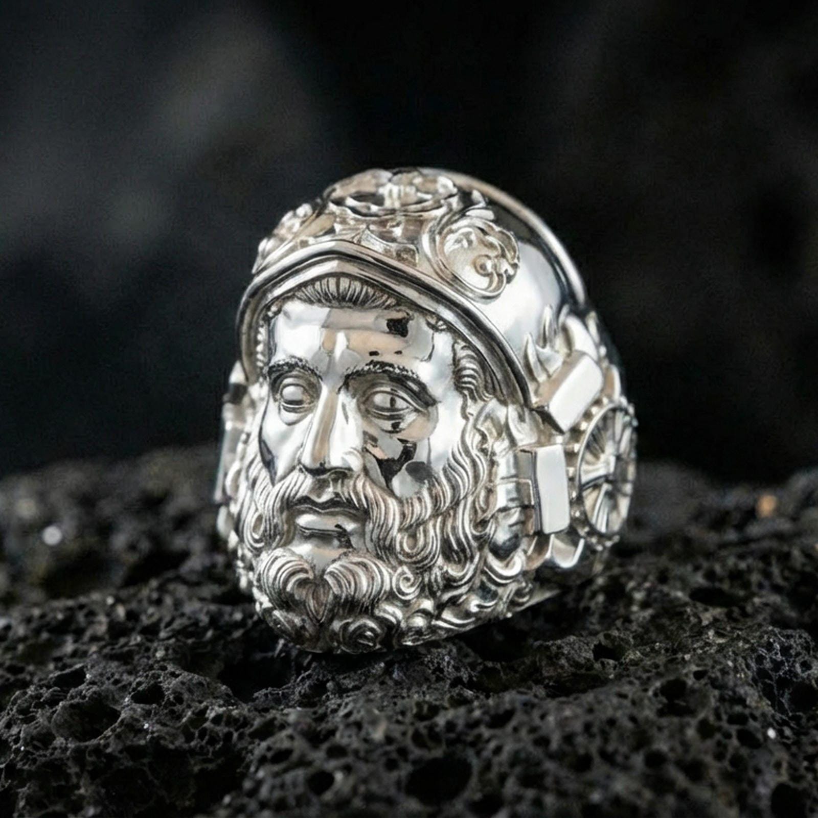 Polished silver Hephaestus ring on volcanic rock, crisp sculpted face design inspired by Greek mythology and craftsmanship