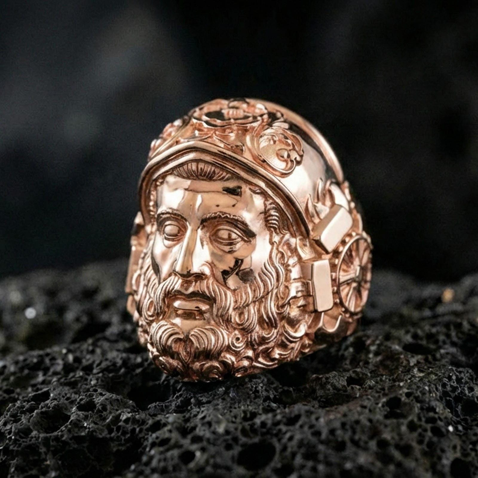 Rose gold plated Hephaestus ring on volcanic rock, myth-inspired signet ring featuring powerful bearded forge god face