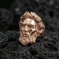 Load image into Gallery viewer, Rose gold plated Zeus ring with high-relief mythology face and laurel wreath detailing, dramatic close-up on black volcanic rock texture
