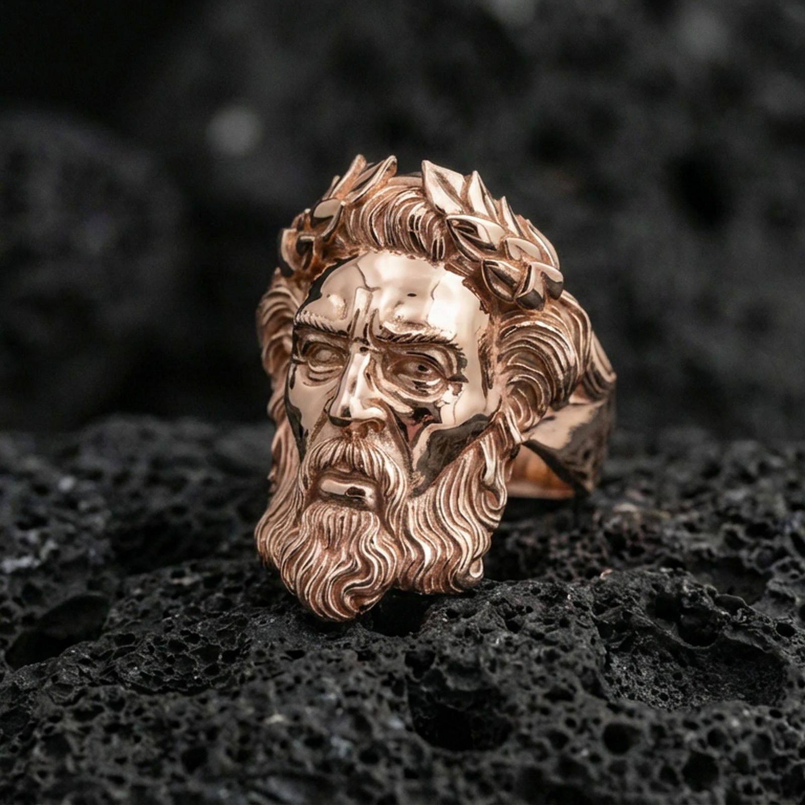 Rose gold plated Zeus ring with high-relief mythology face and laurel wreath detailing, dramatic close-up on black volcanic rock texture