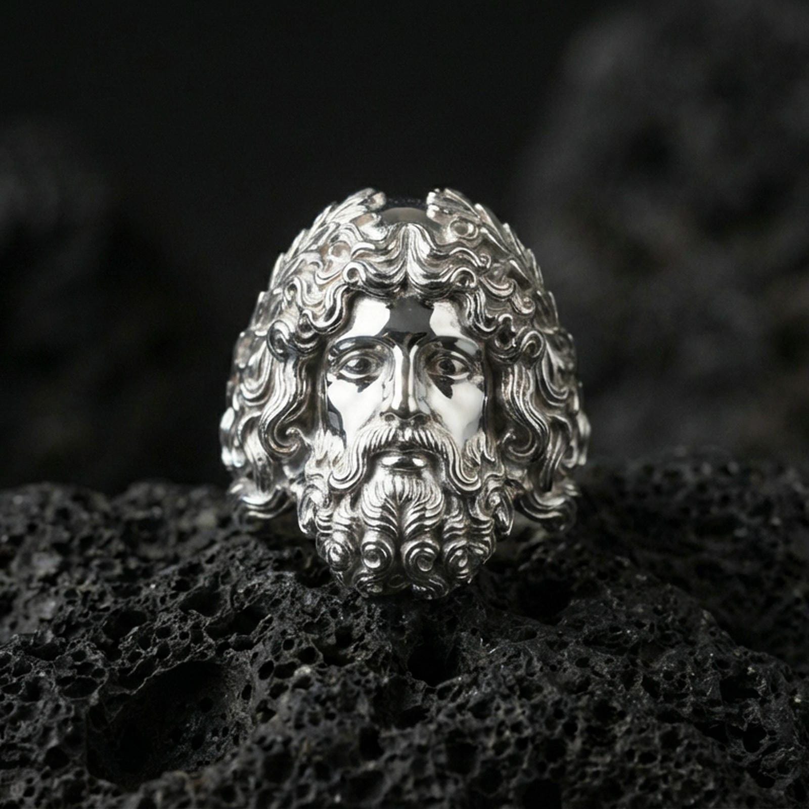 Polished silver Zeus head ring with flowing hair and beard sculpture, centered close-up jewelry shot on dark porous volcanic stone