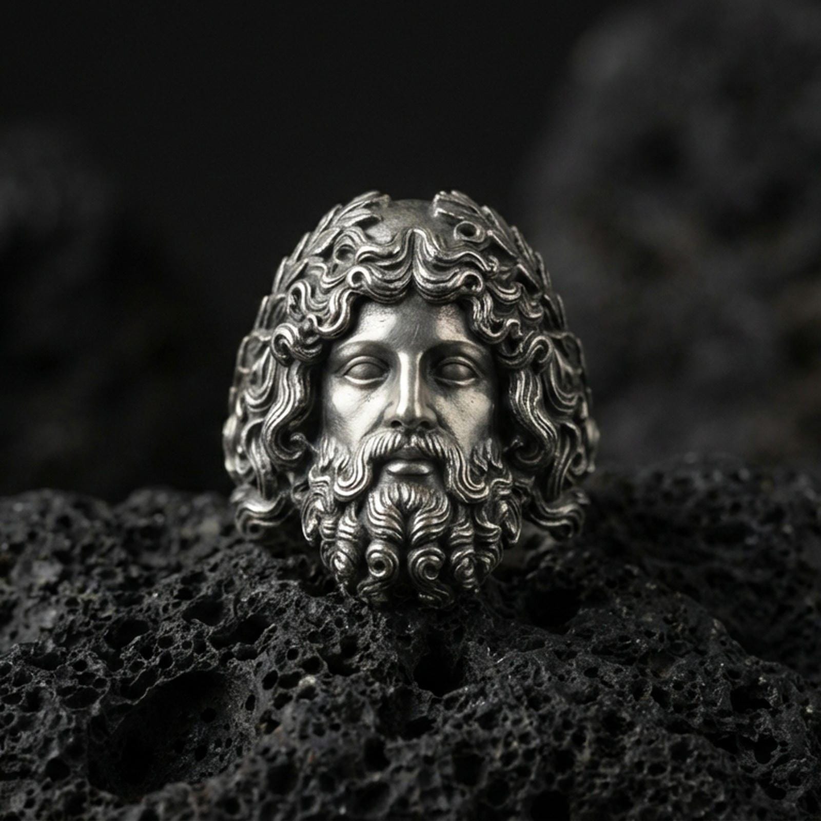 Detailed Zeus head ring crafted in oxidized sterling silver, expressive Greek god facial features, macro product shot on textured volcanic rock background