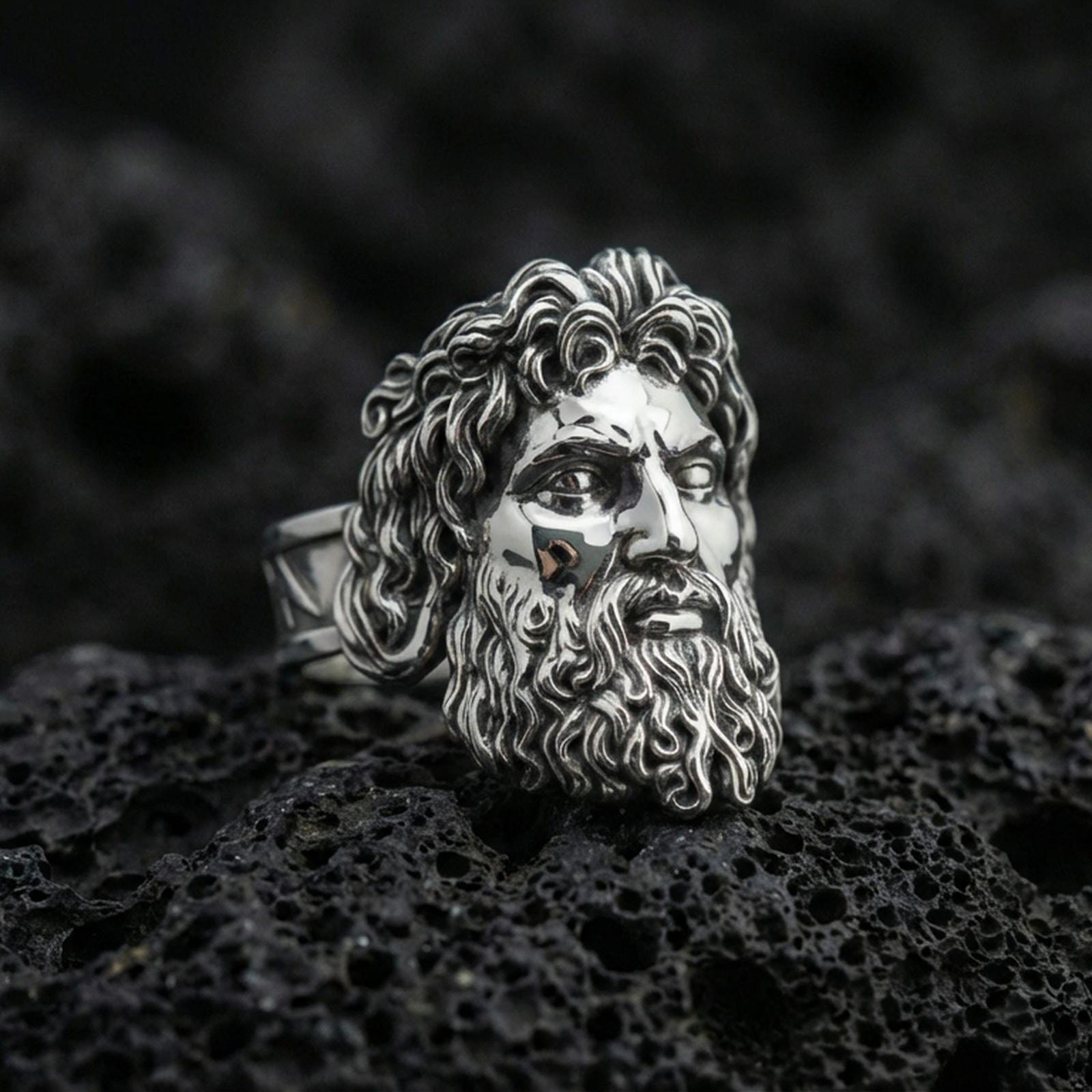 Polished silver Zeus ring with sculpted hair and beard details, centered close-up jewelry photo on dark porous volcanic stone