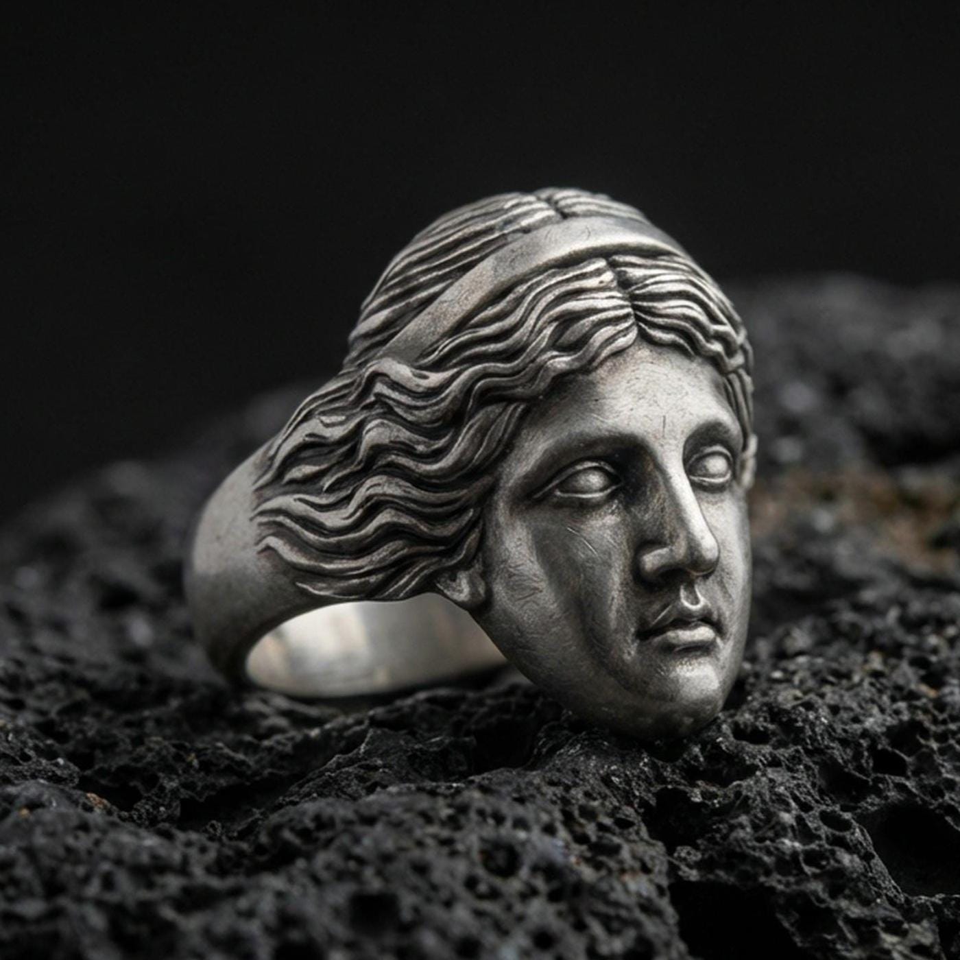 Detailed Venus de Milo ring crafted in oxidized sterling silver, classical Greek goddess face sculpture, macro product shot on textured volcanic rock background