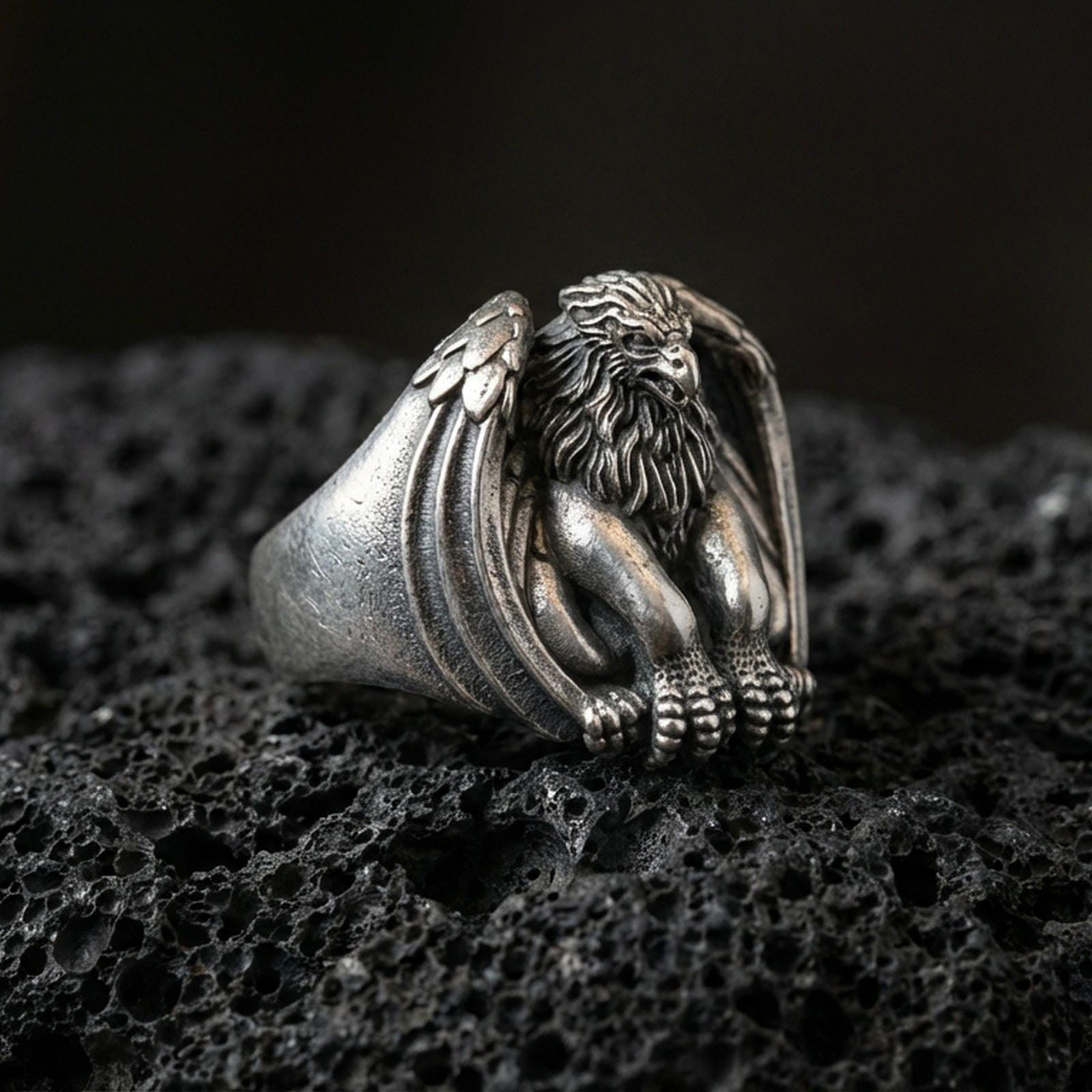 Detailed griffin guardian signet ring crafted in oxidized sterling silver, seated winged beast with lion body, macro product shot on textured volcanic rock background