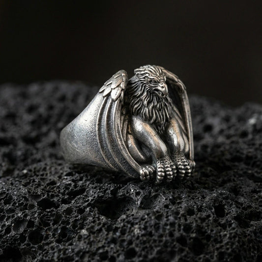 Detailed griffin guardian signet ring crafted in oxidized sterling silver, seated winged beast with lion body, macro product shot on textured volcanic rock background