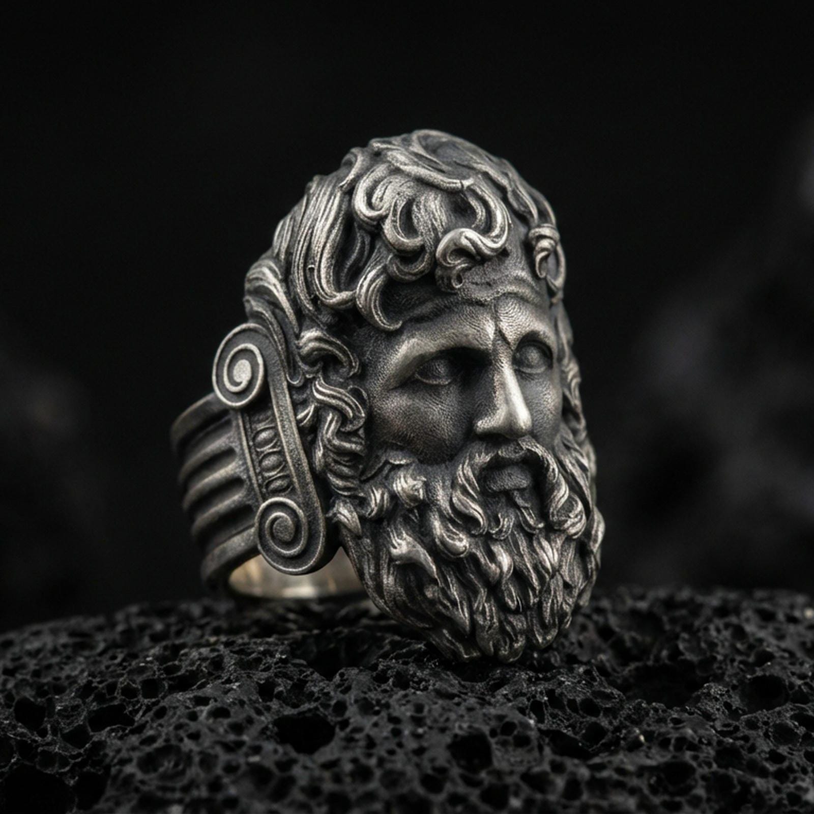 Detailed Zeus ring crafted in oxidized sterling silver, classical Greek god face with flowing beard and ornate curls, macro product shot on textured volcanic rock background